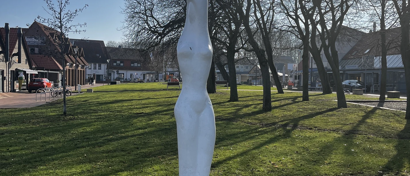 Steinhuder_Nymphe Eine weiße, abstrakte Statue steht auf einem grünen Rasen, umgeben von kahlen Bäumen und Häusern.