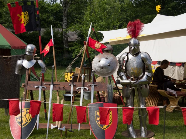 Ritterrüstungen und Schilde auf einem Stand im Mittelaltermarkt Rahden, umgeben von roten Wimpeln.