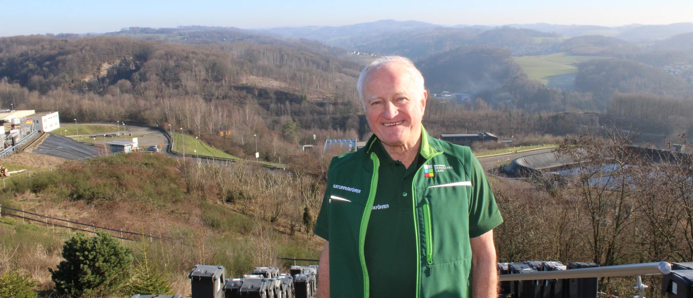Winfried Panske Ein älterer Mann in grüner Kleidung steht lächelnd auf einer Treppe, im Hintergrund hügelige Landschaft.