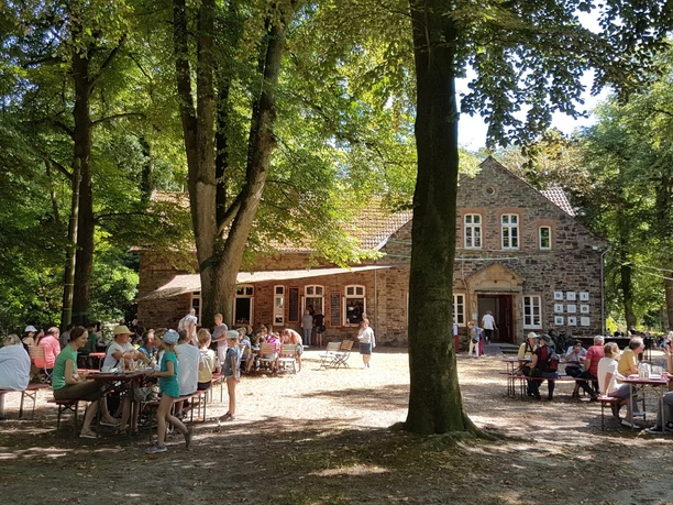 eroeffnung_foto_piesberger_gesellschaftshaus.jpg Menschen sitzen an Tischen im Schatten großer Bäume vor einem historischen Backsteingebäude.