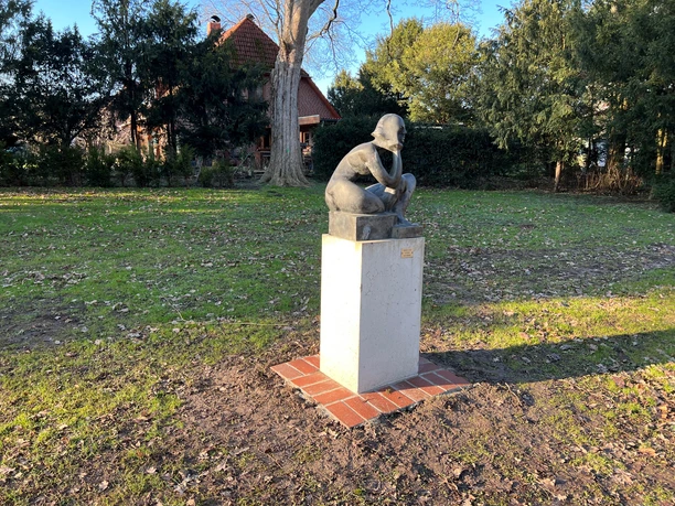 A bronze sculpture of a seated child on a concrete pedestal in a park-like setting.