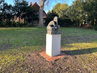 Sitzende Eine Bronzeskulptur eines sitzenden Kindes auf einem Betonsockel in einem parkähnlichen Umfeld.