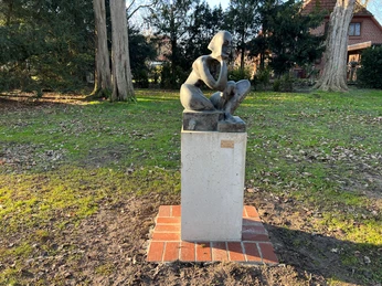 Sitzende2.jpg Skulptur einer hockenden Figur auf einem Sockel, umgeben von Bäumen und Herbstlaub im Freien.