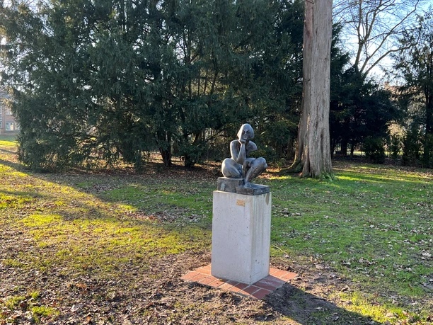 Sitzende Steinskulptur einer nachdenklichen Frau sitzt auf einem Sockel im sonnigen Park mit Bäumen.
