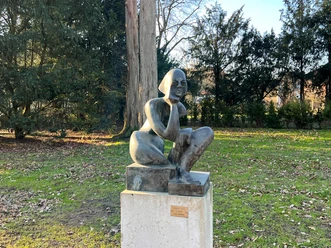 Bronzestatue einer hockenden Frau auf einem Steinpodest in einem parkähnlichen Umfeld aus Bäumen.