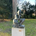 Sitzende Bronzestatue einer hockenden Frau auf einem Steinpodest in einem parkähnlichen Umfeld aus Bäumen.