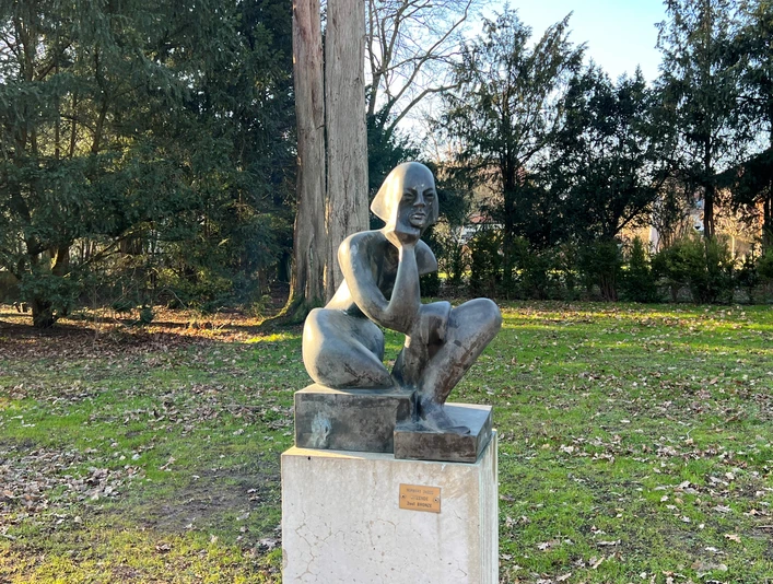 Sitzende Bronzestatue einer hockenden Frau auf einem Steinpodest in einem parkähnlichen Umfeld aus Bäumen.