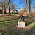 Skulptur einer stilisierten Raubkatze auf einem Sockel in einem sonnigen Park mit alten Gebäuden.