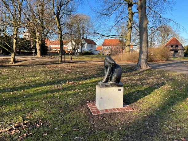 Skulptur einer stilisierten Raubkatze auf einem Sockel in einem sonnigen Park mit alten Gebäuden.