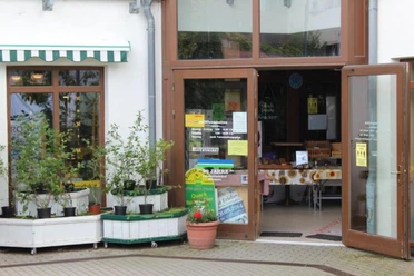 Ingang van een winkel met open deuren, versierd met planten in witte plantenbakken en een tafel met broodjes erin.