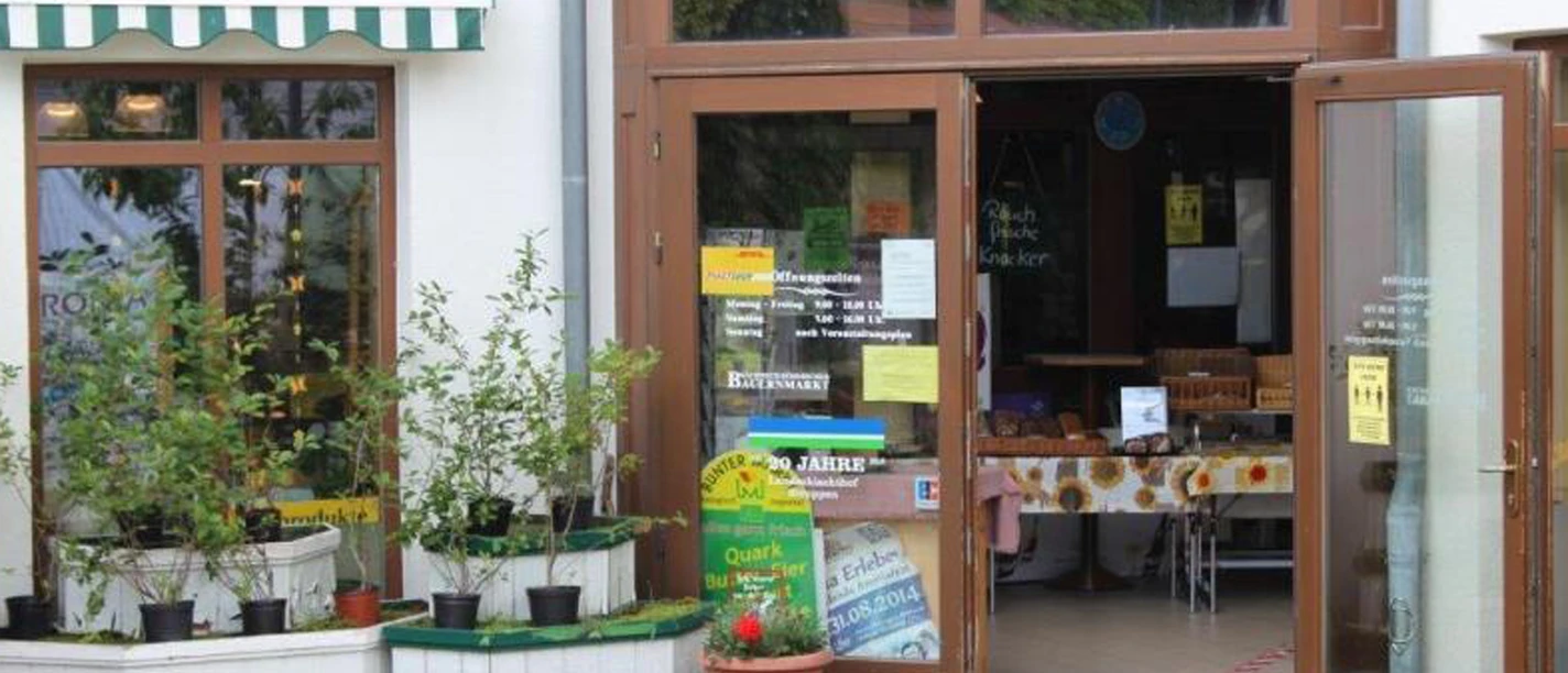 Entrance of a store with open doors, decorated with plants in white planters and a table with sandwiches inside.