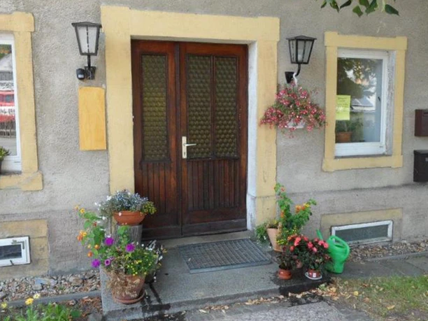 Wooden door with yellow frame, flanked by windows and lanterns; colorful flower pots and green watering can on the floor, inviting atmosphere.