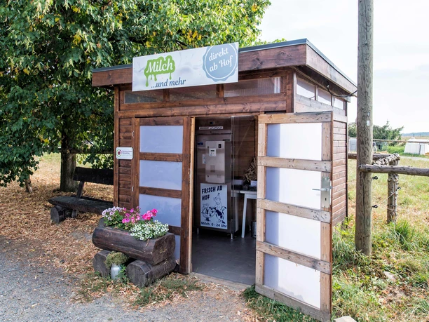 Houten hut met melkautomaat, omringd door bomen en weiland; bordje "Milk ...and more" boven de deur, bloembak met kleurrijke bloemen ervoor.