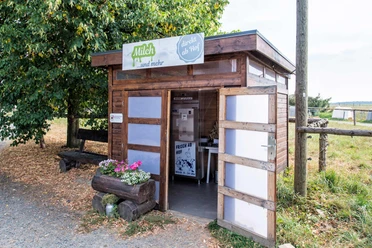 Milchautomat Milchhof Fiedler Holzhütte mit Milchautomat, umgeben von Bäumen und Wiese; Schild "Milch ...und mehr" über der Tür, Blumenkasten mit bunten Blumen davor.