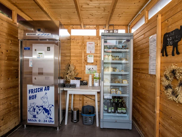 Wooden sales room with milk vending machine on the left and fridge on the right, filled with dairy products. Straw decorations on the walls.