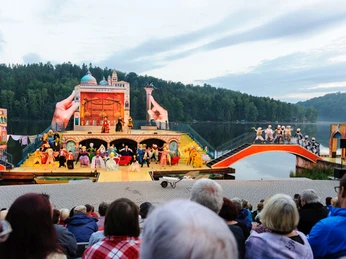 Seebühne Kriebstein_Venedig © Mittelsächsisches Theater - Andre Braun.jpg Freilufttheater vor bewaldetem Seeufer mit Publikum und farbenfroher Bühne.Open-air theater in front of a wooded lakeshore with audience and colorful stage.Divadlo pod širým nebem na břehu zalesněného jezera s publikem a barevnou scénou.Teatr na świeżym powietrzu przed zalesionym brzegiem jeziora z widownią i kolorową sceną.Openluchttheater voor een beboste oever met publiek en een kleurrijk podium.Teatro all'aperto di fronte a una riva boscosa del lago, con un pubblico e un palcoscenico colorato.
