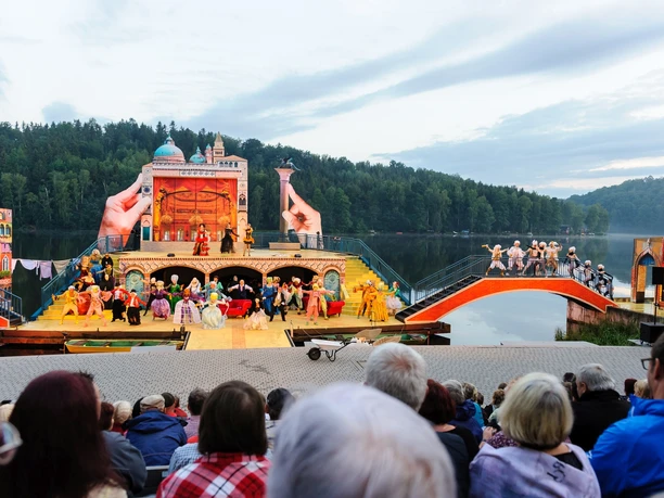 Seebühne Kriebstein_Venedig © Mittelsächsisches Theater - Andre Braun.jpg Freilufttheater vor bewaldetem Seeufer mit Publikum und farbenfroher Bühne.