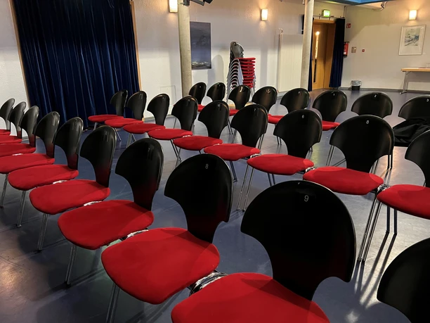 Kulturwerk innen Rote und schwarze Stühle in Reihen, bereit für eine Veranstaltung in einem saalartigen Raum.