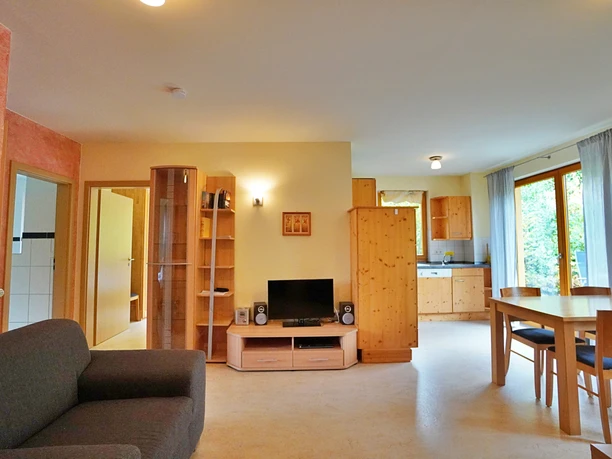 RRA04503-b2.jpg Helles Wohnzimmer mit Sofa, Schrankwand, Esstisch und Zugang zur Küche mit großen Fenstertüren.