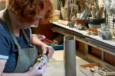 Een persoon met rood haar boetseert klei in een werkplaats; gereedschap en penselen zijn te zien op de achtergrond.