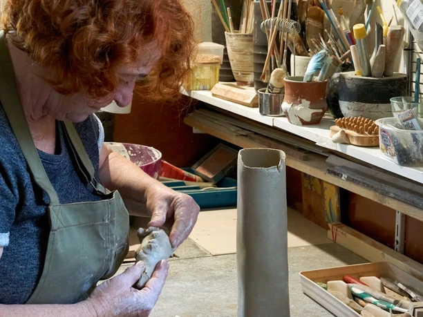 Stefanie Mathy bei der Arbeit im Atelier K Eine Person mit rotem Haar formt Ton in einer Werkstatt; im Hintergrund sind Werkzeuge und Pinsel zu sehen.