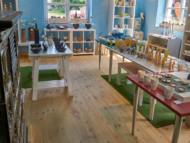 Atelier K in Krippen Ein helles Geschäft mit Holzboden zeigt handgefertigte Keramik und Kunsthandwerk auf weißen Tischen vor blauen Wänden und Regalen.