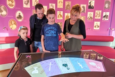 A family looks at an interactive screen in a museum with purple walls and historical portraits.