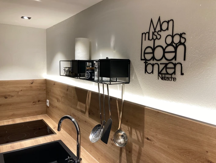 La Petite Maison - Küche 3 In einer modernen Küche hängt ein Zitat an der Wand, Küchenutensilien darunter montiert.