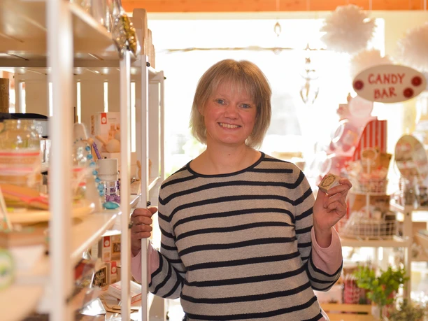 Lächelnde Frau im Hobby-Stübchen, umgeben von Bastelbedarf und Dekoartikeln in Regalen.