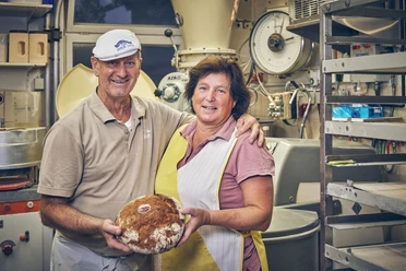 Bäckerei Sand Bäckerpaar mit Brot in der Backstube, Lächeln und umgeben von Backzutaten.
