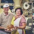 Bäckerei Sand Bäckerpaar mit Brot in der Backstube, Lächeln und umgeben von Backzutaten.