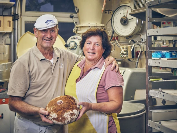 Bäckerei Sand Bäckerpaar mit Brot in der Backstube, Lächeln und umgeben von Backzutaten.