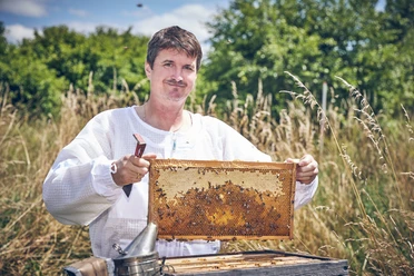Bienenfranz_Spitzenprodukt_Fotomarcgilsdorf.jpg Imker mit Honigwabe, umgeben von Natur, lächelnd in Imkerkleidung.