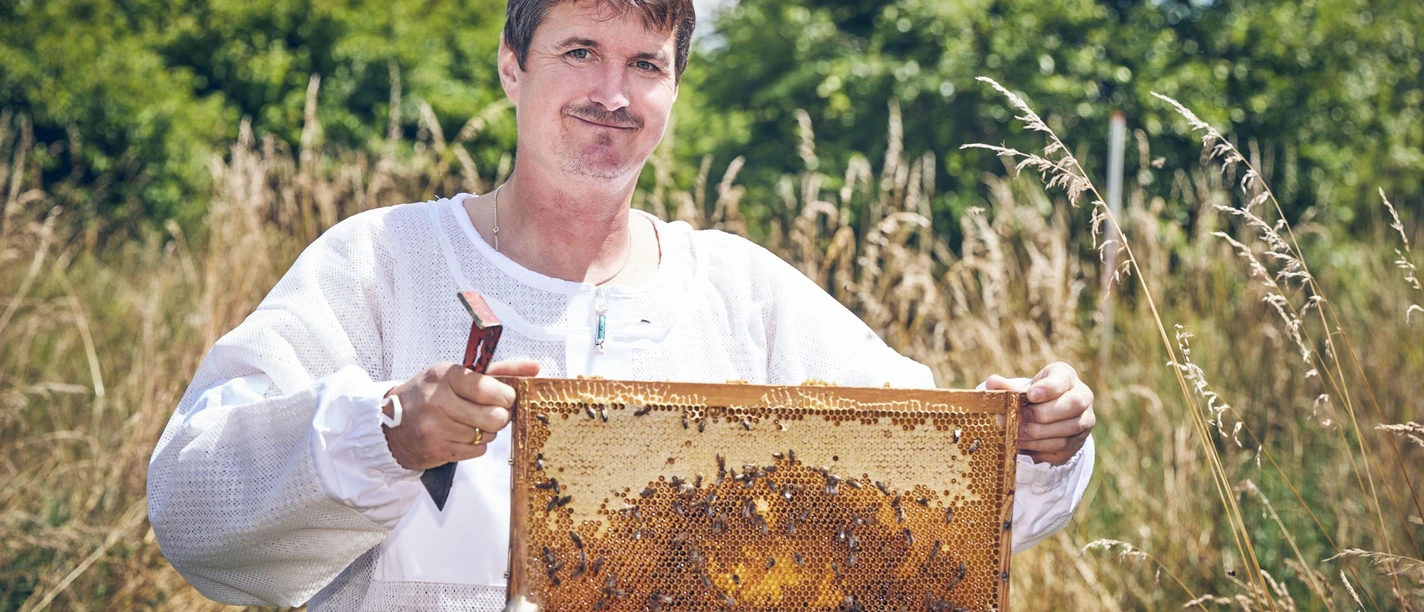 Bienenfranz_Spitzenprodukt_Fotomarcgilsdorf.jpg Imker mit Honigwabe, umgeben von Natur, lächelnd in Imkerkleidung.