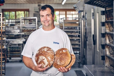 Bäckerei Luidl Mann in Bäckerei hält drei Laibe frisch gebackenes Brot, lächelt in die Kamera.