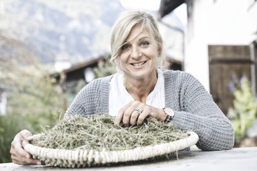Natursula Inser Hoamat Partner Eine lächelnde Frau mit Korb voller Kräuter vor Bergkulisse.