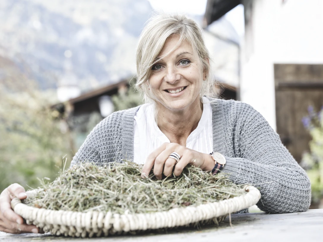 Natursula Inser Hoamat Partner Eine lächelnde Frau mit Korb voller Kräuter vor Bergkulisse.