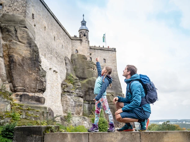 Pevnost Königstein: Muž a dívka v outdoorovém oblečení si prohlížejí velký kamenný hrad; dívka nadšeně ukazuje vzhůru.