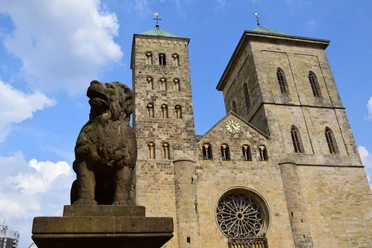 Der sagenhafte Löwenpudel vor dem Osnabrücker Dom Löwenpudel-Skulptur aus Stein mit zwei Türmen des Osnabrücker Doms und blauem Himmel im Hintergrund.