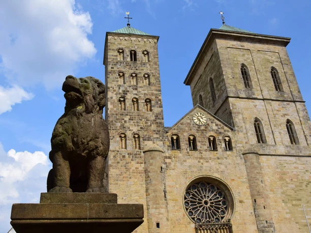 Der sagenhafte Löwenpudel vor dem Osnabrücker Dom Löwenpudel-Skulptur aus Stein mit zwei Türmen des Osnabrücker Doms und blauem Himmel im Hintergrund.
