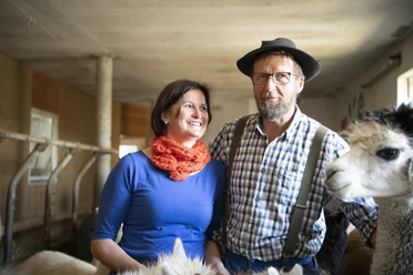 Alpakahof Riegsee Zwei Personen und ein Alpaka in einem Stall stehen nebeneinander.