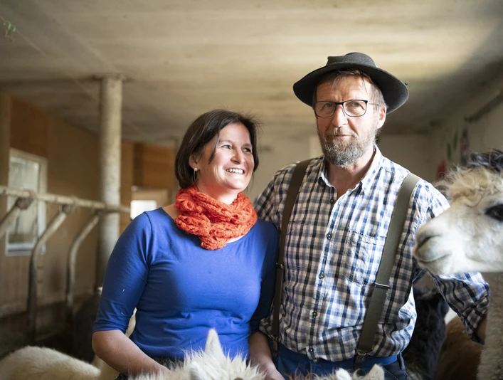 Alpakahof Riegsee Zwei Personen und ein Alpaka in einem Stall stehen nebeneinander.