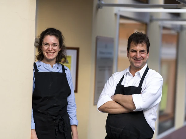 Goldschmiede Thomas Schwabe Eine lächelnde Frau und ein Mann in Schürzen stehen vor einem Gebäude.