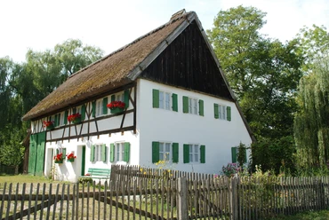 Staudenhaus Oberschönenfeld Weißes Staudenhaus mit grünen Fensterläden und Strohdach, umgeben von blühendem Garten und Holzzaun.