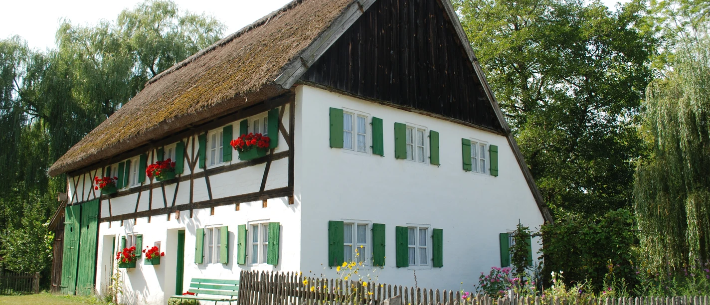 Staudenhaus Oberschönenfeld Weißes Staudenhaus mit grünen Fensterläden und Strohdach, umgeben von blühendem Garten und Holzzaun.