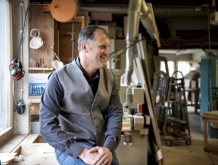 Wohnladen und Schreinerei Geiger Inser Hoamat Partner Ein Mann sitzt in einer Schreinerwerkstatt und lächelt zur Seite.