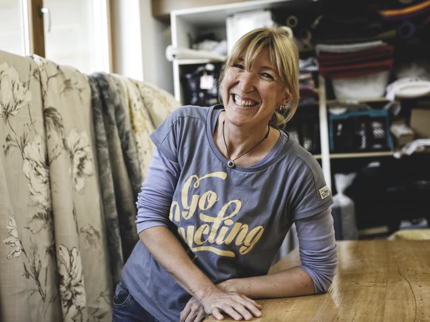 Nöh´s Raumausstattung Inser Hoamat Partner Eine Frau lächelt in einer Werkstatt mit Stoffrollen im Hintergrund.