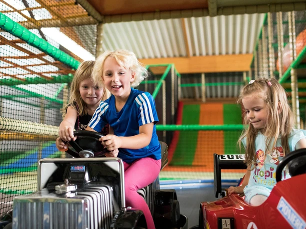 Drei Kinder fahren lachend in kleinen Spielzeugautos in einem Indoor-Spielplatz mit bunten Netzen und Rutschen.