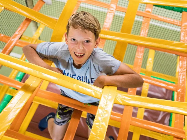 Ein Junge in einem hellblauen T-Shirt klettert lächelnd durch ein gelbes und oranges Klettergerüst in einem Indoor-Spielplatz.