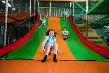 Rutsche im Solivital Sebnitz Vier Kinder rutschen lachend eine große, bunte Rutsche in einem Indoor-Spielplatz hinunter. Die Farben der Rutsche sind grün und orange.